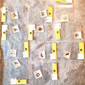 Puede incluir: Una colección de pequeñas tarjetas cuadradas con símbolos del zodiaco en tinta marrón. Las tarjetas están en fundas de plástico transparente con pestañas amarillas y etiquetas blancas. La imagen es desde arriba.