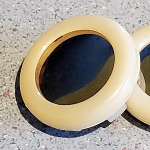 Puede incluir: Primer plano de dos objetos ovalados de color crema con un interior oscuro. Los objetos tienen un acabado brillante y un borde fino de color dorado. Los objetos están sobre una superficie moteada de color gris claro.