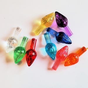 May include: A collection of colorful Christmas light bulbs in various shades, including clear, green, red, orange, yellow, purple, and blue. The bulbs have a classic teardrop shape and are arranged on a white surface.