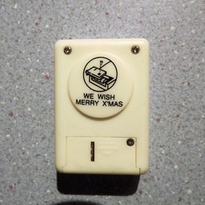 May include: A white plastic Christmas music box with a black and white illustration of a Christmas present. The music box has a button that says "We Wish Merry X'mas".
