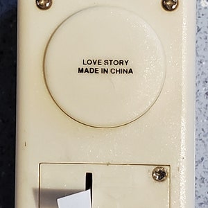 Puede incluir: Una caja de música de plástico blanco con un botón redondo que dice "LOVE STORY MADE IN CHINA". La caja de música tiene un pequeño deslizador blanco en la parte inferior.
