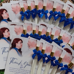 Puede incluir: Recuerdos de boda con peque&ntilde;os frascos de perfume con detalles dorados y lazos azules, dispuestos con tarjetas que dicen "Gracias". Las tarjetas tienen una foto de una pareja y un mensaje de agradecimiento.