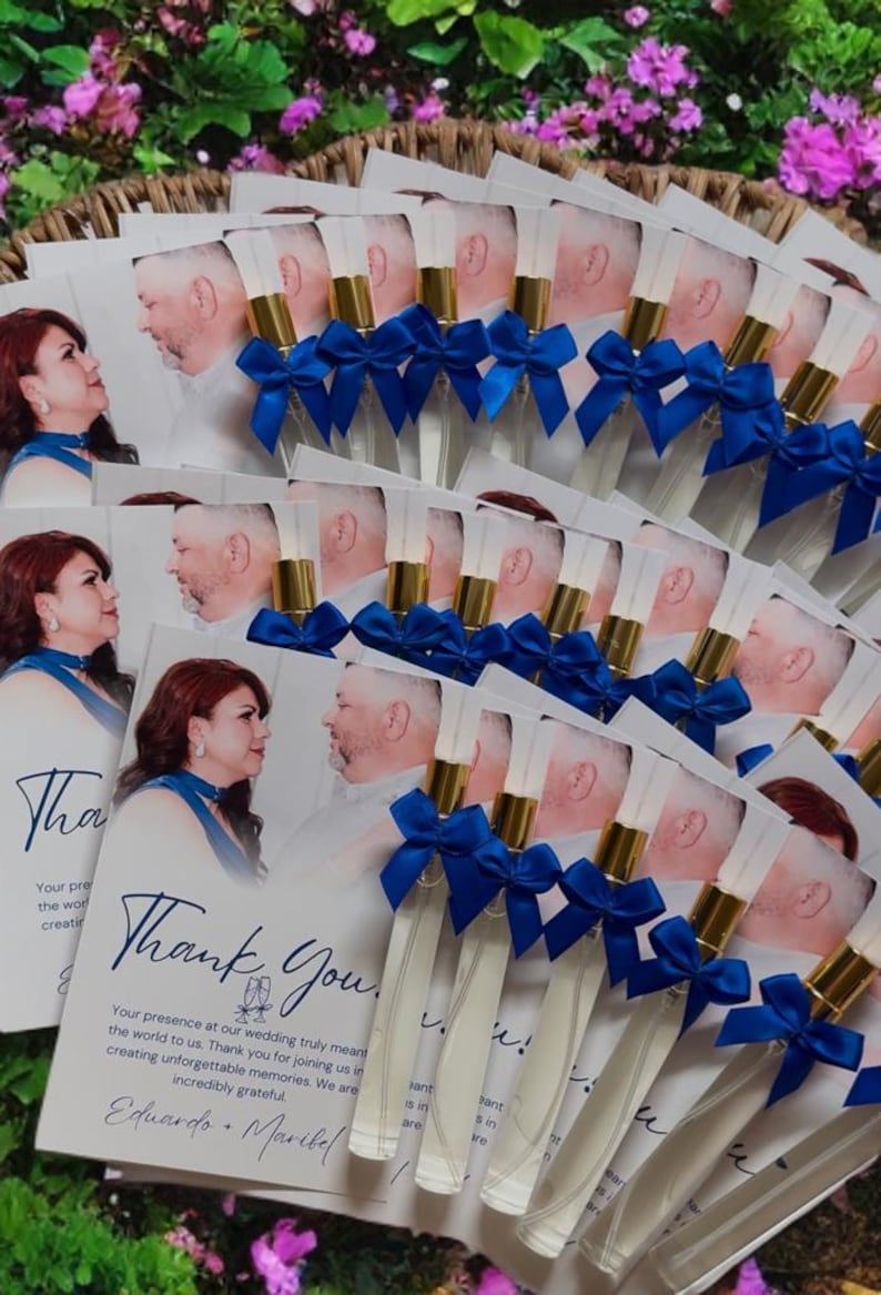 Puede incluir: Recuerdos de boda con peque&ntilde;os frascos de perfume con detalles dorados y lazos azules, dispuestos con tarjetas que dicen "Gracias". Las tarjetas tienen una foto de una pareja y un mensaje de agradecimiento.