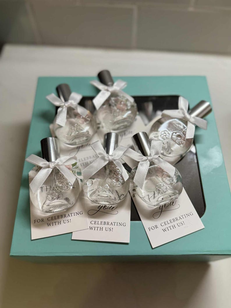 Puede incluir: Un conjunto de seis peque&ntilde;as botellas de vidrio transparente con tapas plateadas, cada una atada con una cinta blanca. Las botellas est&aacute;n en una caja azul claro, con etiquetas que dicen "&iexcl;Gracias por celebrar con nosotros!"