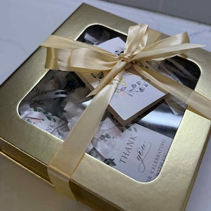 Puede incluir: Una caja de regalo dorada con una ventana transparente, atada con una cinta dorada. La caja contiene art&iacute;culos con dise&ntilde;os florales y etiquetas que dicen "Gracias" y "&iexcl;Por celebrar con nosotros!"