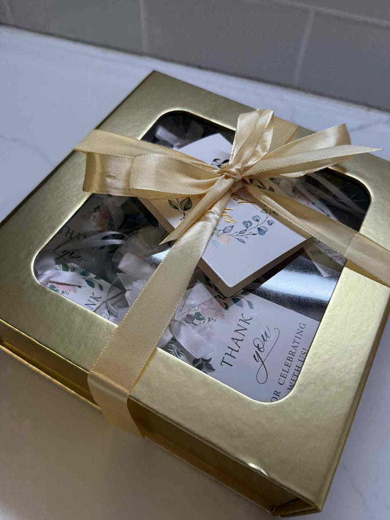 Puede incluir: Una caja de regalo dorada con una ventana transparente, atada con una cinta dorada. La caja contiene art&iacute;culos con dise&ntilde;os florales y etiquetas que dicen "Gracias" y "&iexcl;Por celebrar con nosotros!"