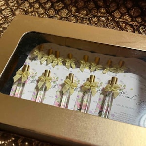 Op de afbeelding: Een gouden doos met kleine glazen flesjes met gouden doppen en strikjes. De flesjes zijn gerangschikt op kaarten met bloemmotieven en de tekst "Thank you". De doos heeft een gouden binnenkant.