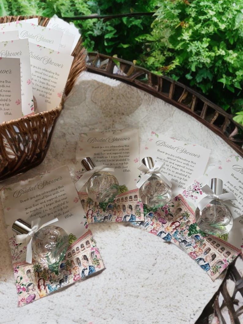 Puede incluir: Recuerdos de despedida de soltera, que incluyen peque&ntilde;os frascos de perfume con tapas plateadas y lazos de cinta blanca, presentados en tarjetas decorativas con ilustraciones florales y arquitect&oacute;nicas. Una cesta de mimbre y vegetaci&oacute;n en el fondo.