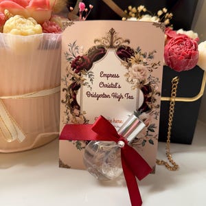 Puede incluir: Un frasco de perfume redondo y transparente con tap&oacute;n plateado y lazo rojo. El frasco est&aacute; frente a una tarjeta rosa con el texto "Empress Christal's Bridgerton High Tea" y dise&ntilde;os florales. Un ramo de flores est&aacute; en el fondo.