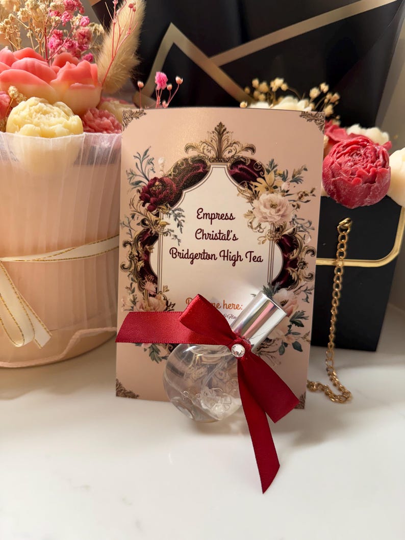 Puede incluir: Un frasco de perfume redondo y transparente con tap&oacute;n plateado y lazo rojo. El frasco est&aacute; frente a una tarjeta rosa con el texto "Empress Christal's Bridgerton High Tea" y dise&ntilde;os florales. Un ramo de flores est&aacute; en el fondo.