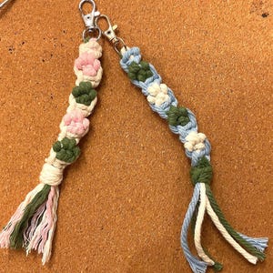 May include: Two macrame keychains with silver-colored clasps. One keychain features pink and green floral designs, while the other has blue, green, and white floral patterns. Both keychains have a tassel of matching colors.