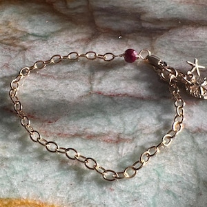 May include: A gold chain bracelet with a red gemstone bead and a charm with a starfish and a heart.