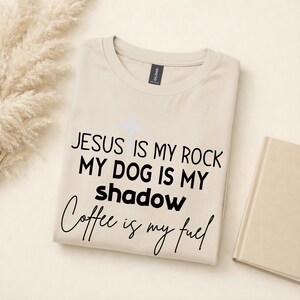 Może przedstawiać: Beżowa koszulka z napisem "JESUS IS MY ROCK, MY DOG IS MY SHADOW, Coffee is my fuel" czarnym pismem. Jasnoniebieski krzyż znajduje się nad tekstem. Z prawej strony złożona książka.