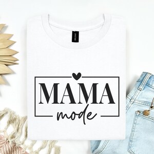 Peut inclure: Un t-shirt blanc à col rond avec une impression graphique noire centrée sur la poitrine. Le motif se compose d'un cadre rectangulaire contenant le texte MAMA dans une police serif grasse et le mot mode en écriture cursive, avec un petit cœur.