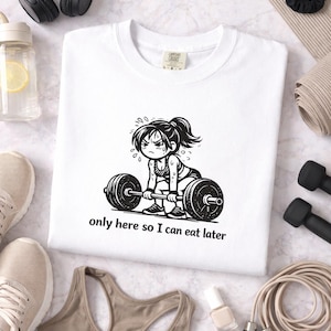Pode incluir: Camiseta branca de gola redonda com um gráfico preto e branco de um levantador de peso e o texto "only here so I can eat later". A imagem inclui acessórios de treino como halteres, uma corda de pular e um sutiã esportivo.