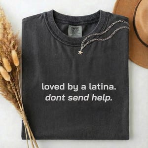 Puede incluir: Camiseta gris oscuro con el texto "loved by a latina. dont send help." Un collar plateado con un colgante de estrella descansa sobre la camiseta. Un sombrero marrón y flores secas están en el fondo.