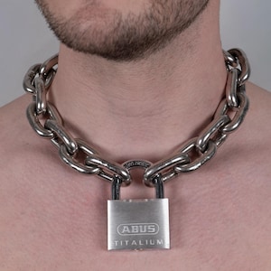 Puede incluir: Un collar de cadena plateada con eslabones grandes y un candado a juego. El candado está grabado con el nombre de la marca "ABUS TITANIUM". El collar se lleva alrededor del cuello.