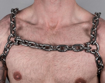May include: A silver-colored chain harness worn across the chest and shoulders. The harness is made of thick, interlocking metal links. The background is a solid gray.