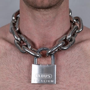 Puede incluir: Un collar de cadena plateada con eslabones grandes rodea el cuello. Un candado plateado con el texto "ABUS TITANIUM" asegura la cadena. El fondo es gris claro liso.