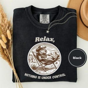 Puede incluir: Una camiseta negra con un gráfico de un capibara con sombrero de vaquero relajándose en una silla de playa. El texto superior dice Relax, y el texto inferior dice NOTHING IS UNDER CONTROL. La camiseta tiene una etiqueta de Comfort Colors.