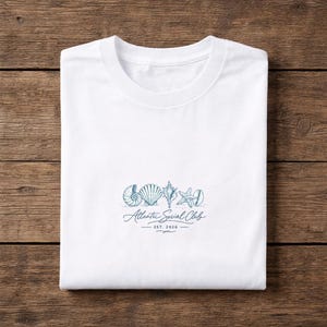 Può includere: T-shirt bianco girocollo con illustrazione blu di conchiglie e la scritta "Atlantic Social Club EST 2026". Il design include un nautilus, una conchiglia, una stella marina e altre creature marine.