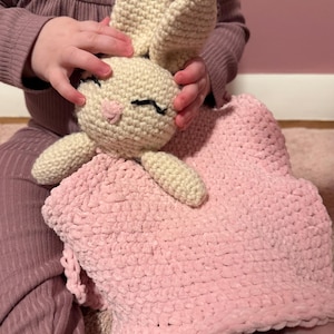 May include: A crochet security blanket featuring a cream-colored bunny head with long ears and embroidered black eyes. The bunny is attached to a soft, textured, light pink square blanket made of plush yarn, creating a cozy and tactile sensory item.