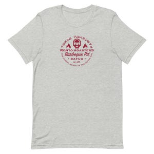 Op de afbeelding: Een lichtgrijs t-shirt met een bordeauxrode cirkelvormige afbeelding. De afbeelding bevat de tekst "RONTO ROASTERS Barbeque Pit" en andere tekst in een gestileerd lettertype. Het t-shirt heeft een ronde hals en korte mouwen.