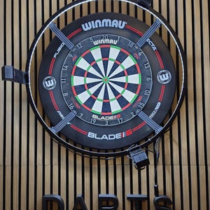 May include: A Winmau Blade 6 dartboard, featuring a black and red design with a white and green bullseye. The board is mounted on a wooden wall, with the word "DARTS" in black letters below. The board has a diameter of approximately 45 cm.