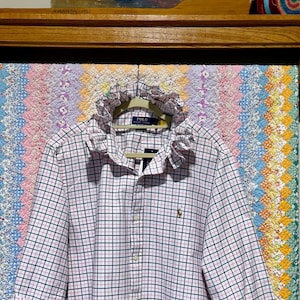 May include: A long-sleeved button-down shirt featuring a white base with a pink and dark green plaid pattern. The shirt includes a ruffled collar, a front button closure, and a small embroidered logo on the left chest area. The cuffs are folded.