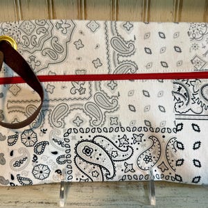 May include: A rectangular zippered pouch featuring a white fabric base with a black and gray paisley bandana print. The pouch includes a bright red horizontal zipper closure and a dark brown leather wrist strap attached by a gold metal ring.