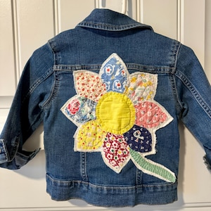 May include: A blue denim jacket with a large, colorful flower applique on the back. The flower features a yellow center and petals made from various patterned fabrics. The jacket has a classic collar and button closures.