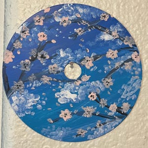 Puede incluir: CD pintado a mano con un diseño floral. La obra de arte representa ramas con flores rosas sobre un fondo de cielo azul. El CD tiene un agujero central y está montado en una pared.