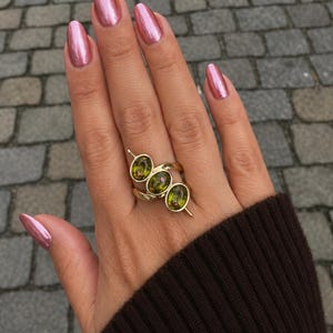 Pode incluir: Um anel de ouro com três pedras ovais de cor verde-oliva dispostas numa fila diagonal. A banda é de ouro polido e as pedras estão cravadas em biséis de ouro individuais, criando um design em camadas com várias pedras no dedo.