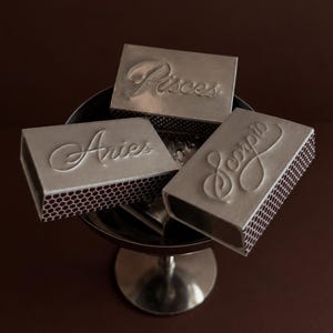 May include: Three rectangular matchboxes with silver-toned covers, each embossed with a different zodiac sign name in cursive script: Aries, Pisces, and Scorpio. The sides of the boxes feature a dark brown honeycomb strike pattern.