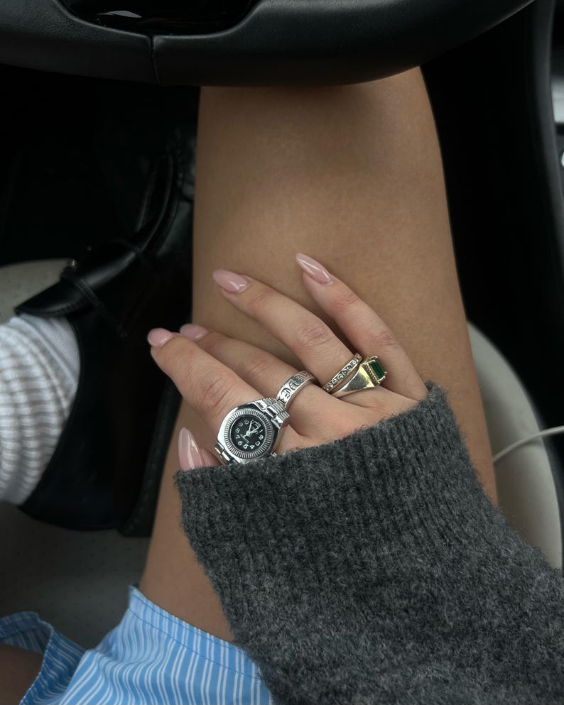 Op de afbeelding: Een hand met meerdere ringen, waaronder een zilveren horlogering en een gouden ring met een groene steen. De hand wordt bedekt door een grijze trui. De nagels zijn lichtroze gelakt.