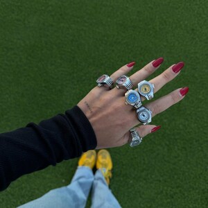 Op de afbeelding: Een hand met meerdere zilveren horlogeringen, elk met een andere gekleurde wijzerplaat. De ringen worden op meerdere vingers gedragen. De achtergrond is groen gras, met de persoon die een blauwe spijkerbroek, gele sneakers en een zwart shirt met lange mouwen draagt.