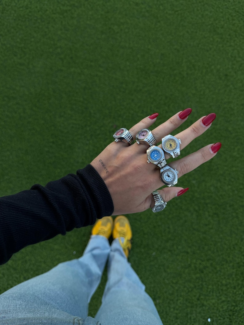 Op de afbeelding: Een hand met meerdere zilveren horlogeringen, elk met een andere gekleurde wijzerplaat. De ringen worden op meerdere vingers gedragen. De achtergrond is groen gras, met de persoon die een blauwe spijkerbroek, gele sneakers en een zwart shirt met lange mouwen draagt.