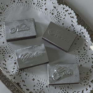 May include: Silver matchboxes with embossed names: Nadia, Sienna, Charlotte, and a box with the word "You". The matchboxes are arranged on a silver tray with a white, lacy paper doily.