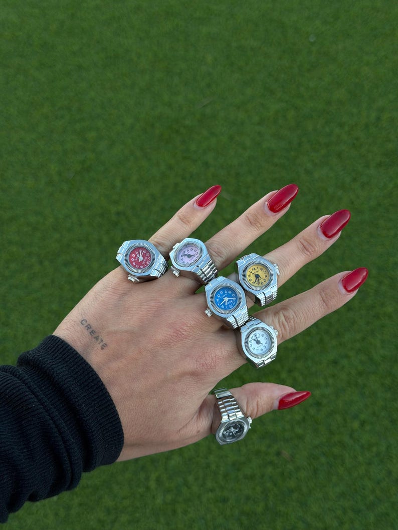Op de afbeelding: Een hand met verschillende zilveren horlogeringen met kleurrijke wijzerplaten. De ringen hebben rode, roze, blauwe en gele wijzerplaten. Het woord "CREATE" is op de pols getatoe&euml;erd. De achtergrond is groen.