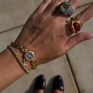 Op de afbeelding: Een verzameling gouden sieraden, waaronder een horloge met een donkerblauwe wijzerplaat en een gouden armband. Twee ringen zijn zichtbaar, een met een zilveren band en gele wijzerplaat, en een andere met een gouden band en een rode steen. Het woord "CREATE" is op de hand getatoe&euml;erd.