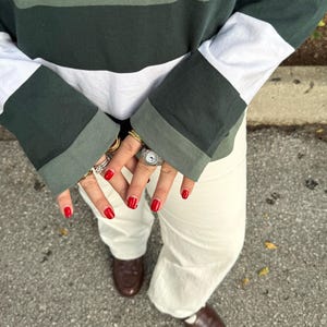Op de afbeelding: Een groen, wit en donkergroen gestreept shirt met lange mouwen, een witte broek en bruine schoenen. De persoon draagt meerdere ringen en heeft rode nagellak.