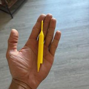 May include: A bright yellow plastic tool, approximately 4 inches long, held in an open hand. The tool has a pointed end and a flat, rectangular body with a small notch. The background shows a wooden floor.