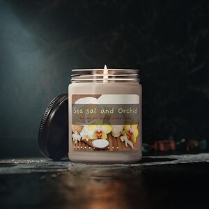 May include: A lit, sea salt and orchid scented candle in a clear glass jar with a silver lid. The label features the text "Sea sal and Orchid" with images of orchids and sea salt. The black lid is off to the side.