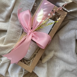 Może przedstawiać: Prostokątne pudełko prezentowe przewiązane różową wstążką i ozdobione suszonymi kwiatami. Wewnątrz pudełka znajduje się świeca i kartka. Na świecy widnieje napis "Lovely House" i "ESS". Pudełko leży na beżowym tle z tkaniny.