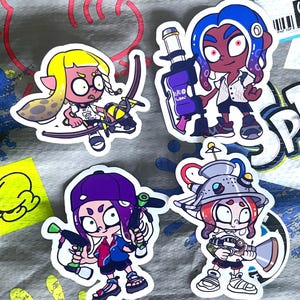 May include: Four colorful stickers featuring characters from the video game Splatoon. Each sticker depicts a character with unique hairstyles, outfits, and weapons. The stickers are on a gray background.