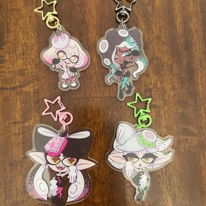 May include: Four acrylic character keychains with star-shaped clasps. The keychains feature colorful anime-style illustrations of characters with unique hairstyles and outfits. The keychains are in pink, white, black, green, and gold.