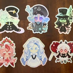 May include: A collection of six holographic stickers featuring cartoon characters in various outfits. The stickers showcase characters in sailor, captain, and witch-like attire, with vibrant colors and glittery accents. The stickers are on a wooden surface.