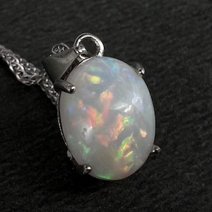 Pode incluir: Um colar com pingente de prata com uma pedra preciosa de opala oval. A opala exibe uma base branca leitosa com flashes iridescentes de cor. O pingente é fixado em uma moldura de prata e pende de uma delicada corrente de prata.