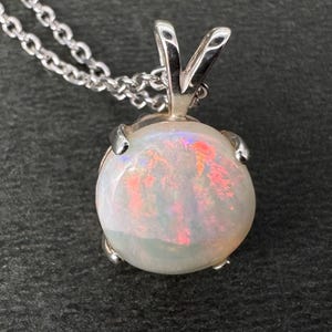 May include: A silver necklace featuring a round opal pendant. The opal has a milky white base with flashes of pink and orange. The pendant is held in place by a silver setting and hangs from a delicate silver chain.