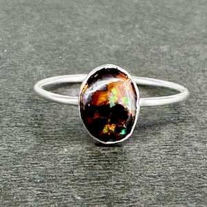May include: A silver ring featuring a polished, oval-shaped ammolite gemstone. The stone displays a dark brown base with iridescent flashes of green, orange, and red. The band is thin, smooth, and circular, holding the stone in a simple bezel setting.